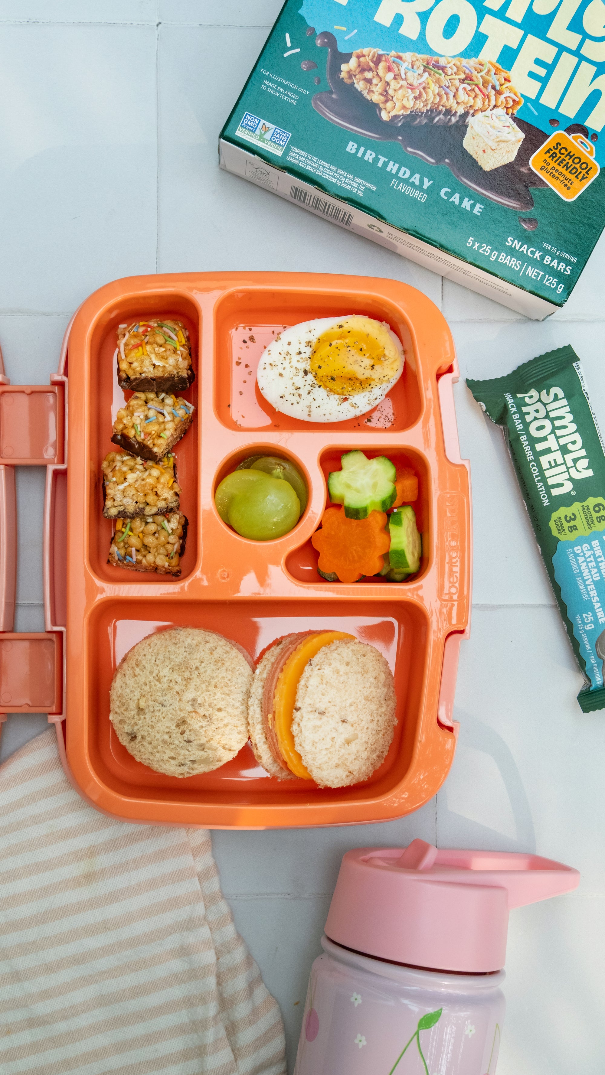 Orange bento box with food compartments on a white surface, including a protein bar and a pink sippy cup.