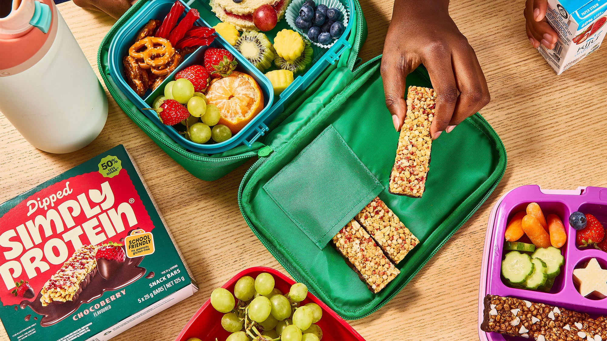 Green bento box with snacks and a hand reaching for a protein bar, surrounded by other bento boxes and protein packaging.