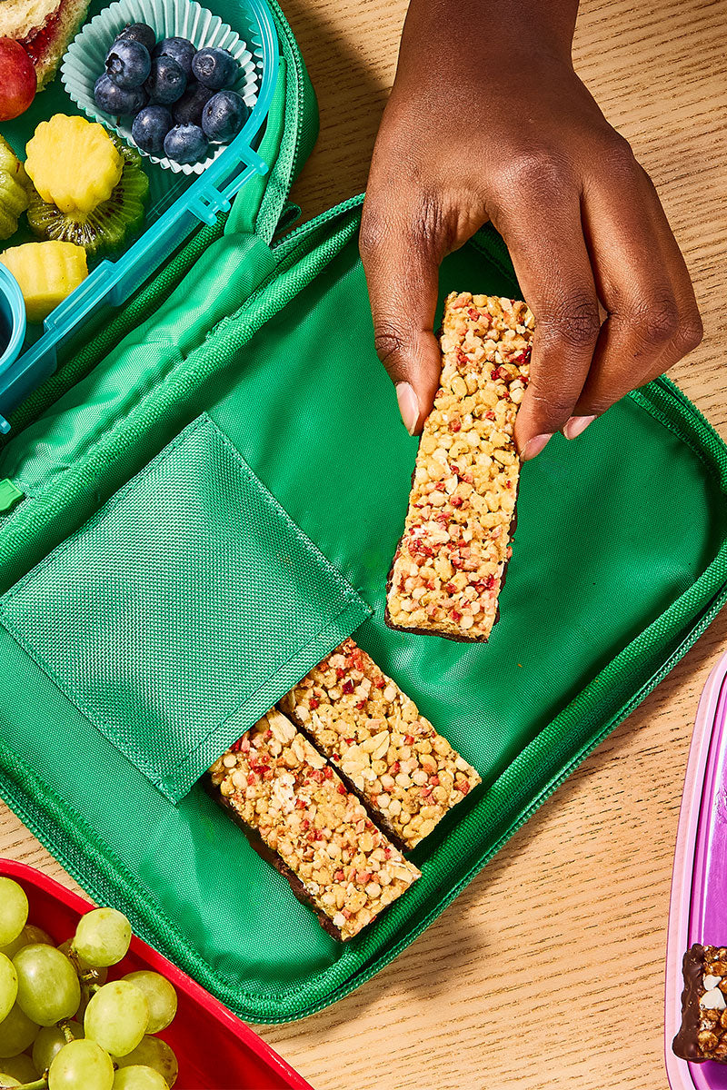 Hand reaching for a granola bar from a green lunchbox with snacks on a wooden surface